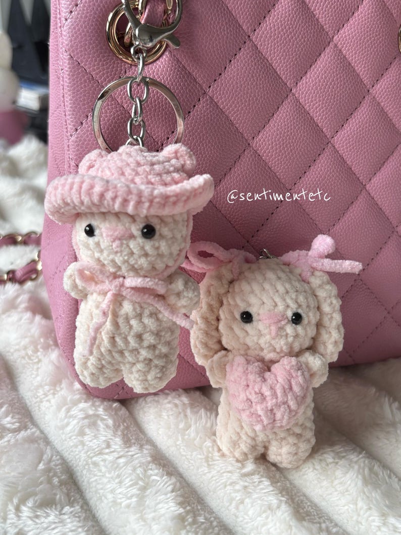 May include: Two handmade crochet bunny keychains. One bunny wears a pink cowboy hat and bow tie, the other holds a pink heart. Both are cream-colored with pink accents. The background includes a pink quilted purse and a fluffy white blanket.