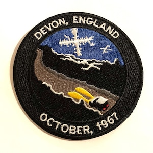 May include: Black and white embroidered patch with a blue and white snowflake, a dark blue sky with white clouds, a gray and black mountain range, and a car with yellow headlights. The text on the patch reads "DEVON, ENGLAND OCTOBER, 1967".