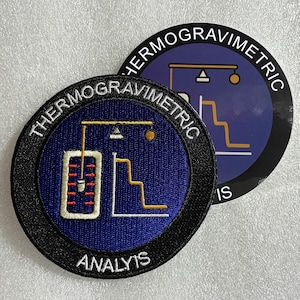 May include: A circular embroidered patch and a matching circular sticker featuring a blue background with a white border. The design includes a yellow and white graphic of a laboratory scale and a step graph, with the text THERMOGRAVIMETRIC ANALYSIS.