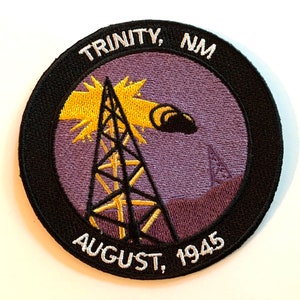 May include: A black and white embroidered patch with a purple background. The patch features a stylized image of a radio tower with a yellow explosion above it. The text "TRINITY, NM AUGUST, 1945" is embroidered around the edge of the patch.