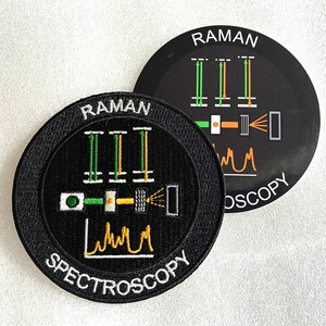 May include: A circular black embroidered patch and a matching sticker featuring the text RAMAN SPECTROSCOPY. The design displays a scientific diagram with green and orange vertical lines, a horizontal sequence of shapes, and a yellow line graph.