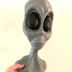 May include: A gray alien head sculpture with large black eyes. The alien has a smooth, rounded head and a slender neck. The sculpture is held by a hand, and the background is a plain, off-white wall.