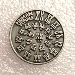 Puede incluir: Un pin de metal plateado con un diseño circular que representa el cariotipo femenino humano, mostrando 23 pares de cromosomas, incluyendo el par XX. El pin está etiquetado como "HUMAN FEMALE" en la parte superior.
