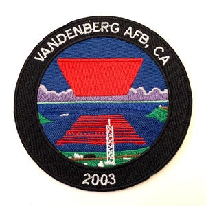 Może przedstawiać: Naszywka haftowana w kolorze czarnym i białym z tekstem "VANDENBERG AFB, CA 2003". Naszywka przedstawia czerwony i niebieski wzór z wystrzeleniem rakiety.