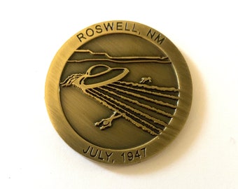 May include: A gold-coloured metal coin featuring a depiction of a flying saucer landing in a field. The coin is inscribed with the text "ROSWELL, NM" and "JULY, 1947".