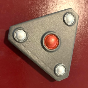 May include: A gray, triangular object with three silver-colored circular accents and a central, raised, red-orange button. The object has a textured surface and is set against a red background. The design is geometric and modern.