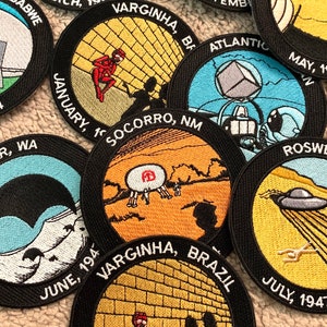 May include: A collection of embroidered patches with black borders. Each patch features a unique design with text, including dates and locations like Roswell, NM, and Varginha, Brazil. The patches depict UFOs, aliens, and other related imagery.
