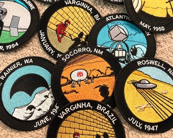 Peut inclure: Plusieurs écussons brodés avec des bordures noires. Chaque écusson présente un design unique avec du texte, notamment des dates et des lieux comme Roswell, NM, et Varginha, Brésil. Les écussons représentent des ovnis, des extraterrestres et des images connexes.