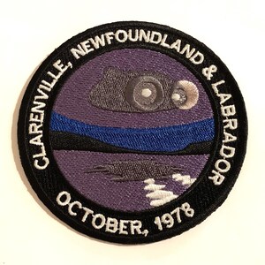 Puede incluir: Un parche bordado en blanco y negro con el texto "Clarenville, Terranova y Labrador Octubre 1978" alrededor de un diseño morado y azul.