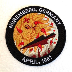 May include: Embroidered patch with a black background and white text that reads "Nuremberg, Germany April, 1561". The center of the patch features a yellow sun with a red cross and red dots, and a red and black spear.
