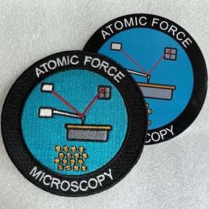 May include: Two circular patches featuring the text ATOMIC FORCE MICROSCOPY. The design includes a blue background, a black border, and a graphic illustration of a cantilever probe scanning a surface with a red laser beam and gold atoms.