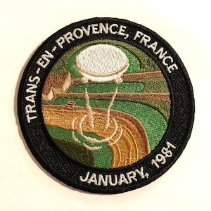 May include: A black and white embroidered patch with a green and gold design. The patch features a white dome with a black outline and the text "TRANS-EN-PROVENCE, FRANCE JANUARY, 1981".