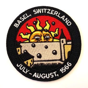 Op de afbeelding: Zwart-witte geborduurde patch met een rode, gele en witte zon met een gezicht en een wit kasteel op de achtergrond. De tekst "BASEL, SWITZERLAND JULY - AUGUST, 1566" staat rond de rand van de patch.