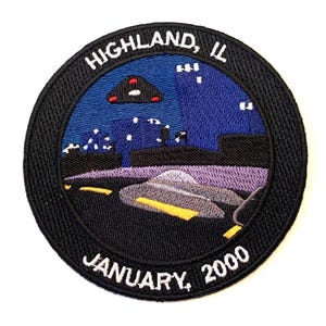 May include: Embroidered patch with a black background and a white circle containing a cityscape with a UFO flying over it. The text "HIGHLAND, IL JANUARY, 2000" is written around the circle.