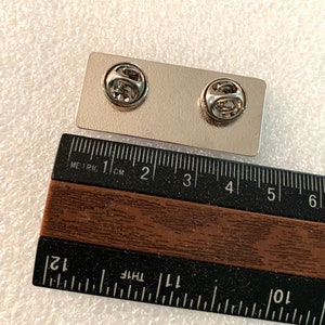 May include: A silver metal pin with a rectangular backing. The pin is approximately 5 cm long and 2.5 cm wide. The pin has a safety clasp on the back.