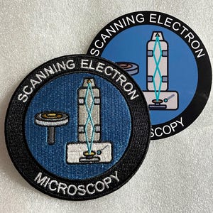 Peut inclure: Deux écussons circulaires présentant un centre bleu avec une illustration graphique d'un microscope électronique à balayage et le texte SCANNING ELECTRON MICROSCOPY en lettres blanches sur la bordure noire. Le design montre une colonne.
