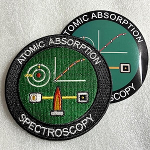 Peut inclure: Un écusson brodé circulaire et un autocollant assorti sur fond vert avec le texte ATOMIC ABSORPTION SPECTROSCOPY. Le motif comprend un modèle atomique blanc, une icône de flamme rouge et orange, et un graphique linéaire blanc avec des points rouges.