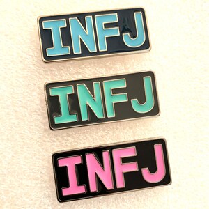 May include: Three enamel pins with the letters "INFJ" in black with different coloured backgrounds. The top pin has a light blue background, the middle pin has a light green background, and the bottom pin has a pink background.