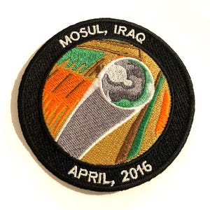 Può includere: Una toppa rotonda ricamata in bianco e nero con il testo "Mosul, Iraq Aprile 2016" e un'immagine stilizzata di un edificio con una finestra.