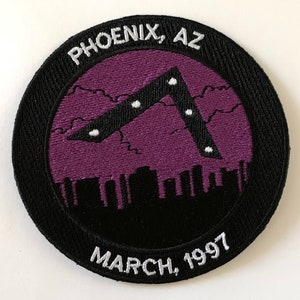 May include: A black and purple embroidered patch with a white outline. The patch depicts a dark, triangular shaped object with white lights in the sky over a city skyline. The text "PHOENIX, AZ" and "MARCH, 1997" are embroidered in white on the patch.