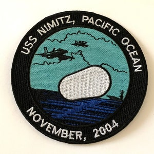 May include: A black and white embroidered patch with the text "USS Nimitz, Pacific Ocean, November, 2004". The patch features a white cloud and two fighter jets flying over a blue ocean.