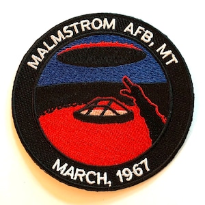 May include: Embroidered patch with a circular design featuring the text "MALMSTROM AFB, MT" and "MARCH, 1967". The patch depicts a red and black landscape with a blue sky and a stylized UFO. A black border surrounds the design.