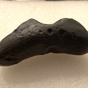 Puede incluir: Un meteorito de color gris oscuro, de forma irregular, con una superficie texturizada. El meteorito tiene un aspecto liso, casi pulido.