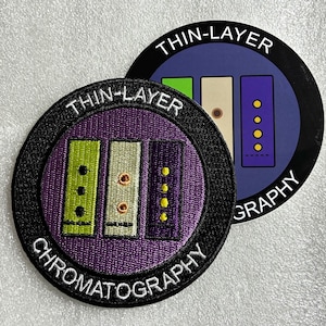 May include: A circular embroidered patch and a matching circular sticker featuring the text THIN-LAYER CHROMATOGRAPHY. Both items display a purple background with three vertical rectangular bars in lime green, beige, and dark purple with dots.