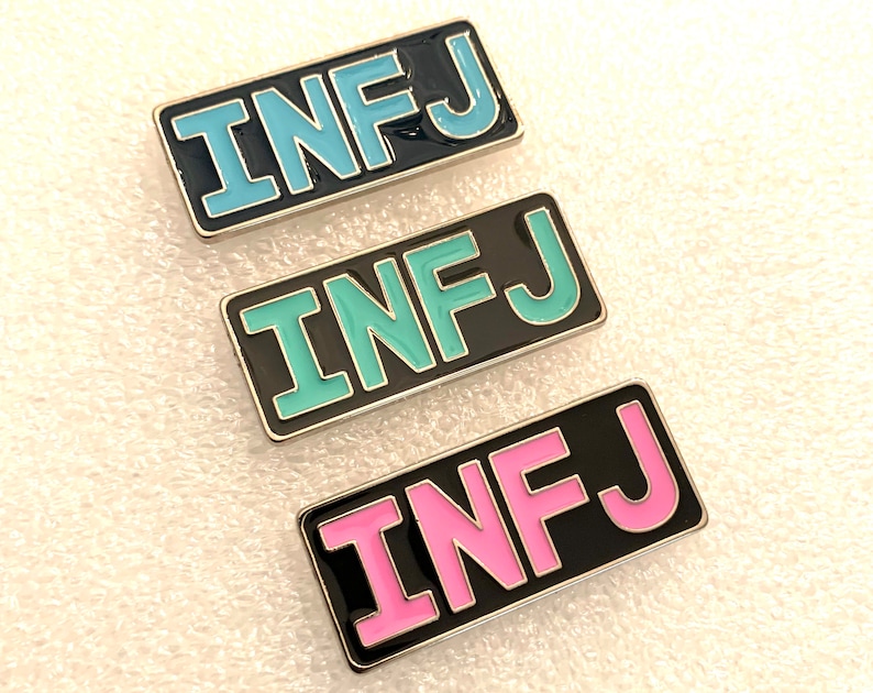 May include: Three enamel pins with the letters "INFJ" in different colours. The top pin is blue, the middle pin is teal, and the bottom pin is pink.