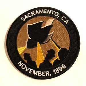 May include: Circular embroidered patch with a black border. The patch features the text "SACRAMENTO, CA" and "NOVEMBER, 1896". It has a brown background with a black silhouette and yellow accents.
