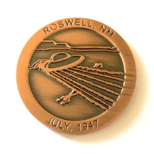 Op de afbeelding: Een koperkleurige munt met een reliëfbeeld van een vliegende schotel en de tekst "ROSWELL, NM JULY, 1947".
