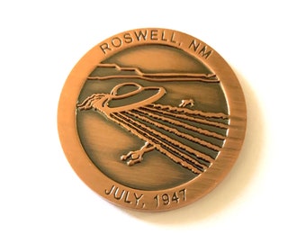 May include: A copper-coloured coin with a raised image of a flying disc and the text "ROSWELL, NM JULY, 1947".