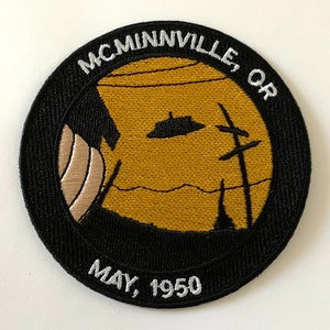 May include: A black and white embroidered patch with the text "MCMINNVILLE, OR MAY, 1950" around the edge. The center of the patch features a yellow silhouette of a landscape with a flying saucer.
