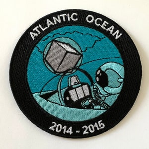 Puede incluir: Un parche bordado en blanco y negro con el texto "ATLANTIC OCEAN" y "2014 - 2015". El parche presenta un astronauta de dibujos animados con un traje espacial azul que sostiene un cubo gris.