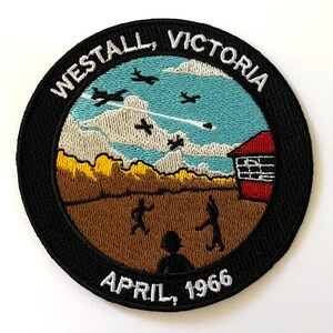 May include: Embroidered patch with a black background and white text that reads "Westall, Victoria April, 1966". The patch depicts a scene of a small town with a red building, a brown field, and a blue sky with white clouds and airplanes.