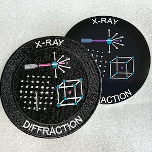 Peut inclure: Deux écussons circulaires noirs avec le texte blanc X-RAY DIFFRACTION. Le motif représente un faisceau frappant une structure cristalline, illustrée par un cube avec des points aux sommets et un point central, ainsi qu'un motif en grille.