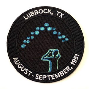 Puede incluir: Parche bordado en blanco y negro con una silueta azul y verde de una persona mirando hacia arriba a una fila de puntos azules y verdes. El texto "LUBBOCK, TX AUGUST - SEPTEMBER 1951" está bordado alrededor del borde.