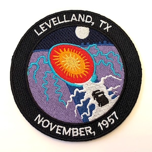 Puede incluir: Parche bordado con un diseño colorido de un sol, una luna y rayos de luz sobre un fondo morado. El texto "LEVELLAND, TX NOVEMBER, 1957" está bordado alrededor del borde del parche.