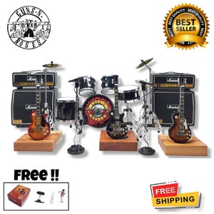 Könnte beinhalten: Eine Miniatur-Nachbildung der Band-Ausrüstung von Guns N' Roses, bestehend aus zwei E-Gitarren, einem Schlagzeug und zwei Verstärkern. Die Gitarren sind braun und schwarz, die Trommeln sind schwarz und silber, und die Verstärker sind schwarz mit dem aufgedruckten Wort "Marshall". Das Schlagzeug hat ein Guns N' Roses-Logo auf der Bassdrum. Das Set wird auf Holzsockeln präsentiert.