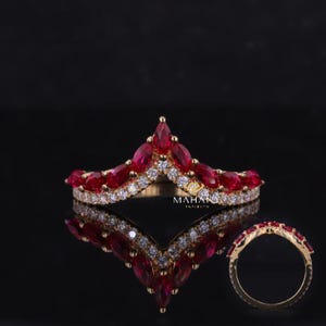 Op de afbeelding: Een gouden ring met een kroonontwerp, met marquise-geslepen rode edelstenen en kleine witte edelstenen. De ring staat tegen een zwarte achtergrond, met een weerspiegeling eronder. Het woord "MAHANA" is zichtbaar.