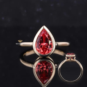 May include: A gold ring featuring a pear-shaped, vibrant red gemstone. The gemstone is set in a bezel setting, and the ring is displayed on a reflective black surface. The ring is shown from multiple angles, highlighting its design and details.