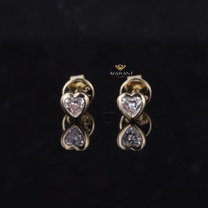 May include: A pair of heart-shaped stud earrings featuring a clear, sparkling stone set in a gold bezel. The earrings are presented on a reflective black surface. The logo "MAHANT" is visible in the upper right corner.