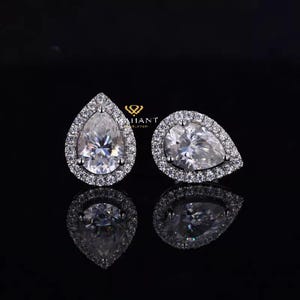 May include: A pair of pear-shaped diamond earrings with a halo setting. The earrings are set in silver and have a white diamond center stone.