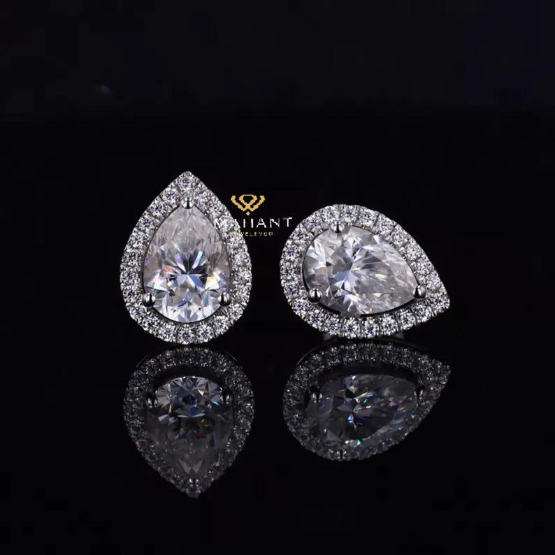 May include: A pair of pear-shaped diamond earrings with a halo setting. The earrings are set in silver and have a white diamond center stone.