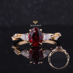 May include: A gold ring featuring a large oval-cut, deep red gemstone as the centerpiece, flanked by two pear-shaped white gemstones. The band has a delicate, leaf-like design. A smaller version of the ring is also visible.