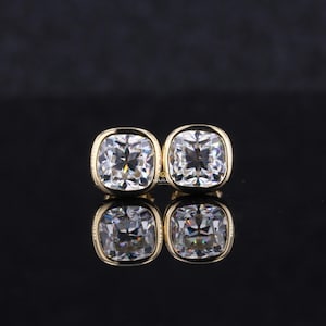 May include: Four square-cut, clear gemstone stud earrings with gold-colored bezels. The earrings are displayed on a reflective black surface, with their reflections visible below. The gemstones have multiple facets, reflecting light.