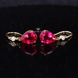 May include: A pair of gold earrings with a teardrop-shaped, vibrant red gemstone. Each earring features a small, round, clear stone and a curved gold wire design. The earrings are displayed on a reflective black surface.