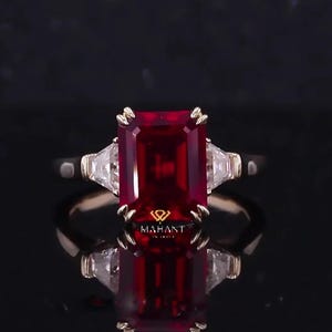 Könnte beinhalten: Ein goldener Ring mit einem großen, rechteckigen, tiefroten Edelstein, flankiert von zwei kleineren, dreieckigen, klaren Edelsteinen. Der Ring hat ein goldenes Band und ist vor einem dunklen Hintergrund platziert. Der Ring hat den Text "Mahant".