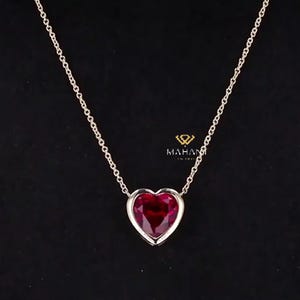 May include: A silver necklace featuring a heart-shaped, deep red gemstone pendant. The pendant is set in a silver bezel and hangs from a delicate chain. The background is black.