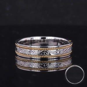 May include: A silver-toned band ring featuring a central engraved scroll pattern bordered by two thin, twisted gold-toned rope edges. The ring has a polished finish and a small logo mark is visible on the inner surface of the band.